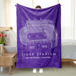 Puede incluir: Una manta de estadio morada con un dibujo arquitectónico blanco del Tiger Stadium en Baton Rouge, Luisiana. El nombre y la ubicación del estadio están impresos en blanco. La manta está sujeta por una persona.