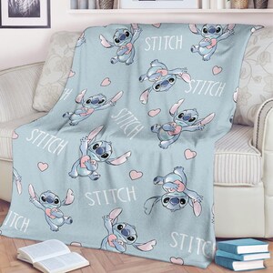 May include: A light blue throw blanket featuring a repeating pattern of the character Stitch holding a heart, with the word "Stitch" and pink hearts scattered across the surface. The blanket is draped over a sofa.