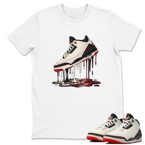 May include: White t-shirt with a graphic of a white and black trainer with red detailing, depicted with dripping black and red paint. A pair of matching trainers are shown at the bottom. The shirt is a casual wear item.