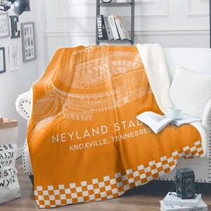 May include: An orange and white stadium blanket featuring a detailed architectural drawing of Neyland Stadium in Knoxville, Tennessee. The blanket has a white checkered border and a soft, white sherpa lining. The blanket is draped over a white couch.
