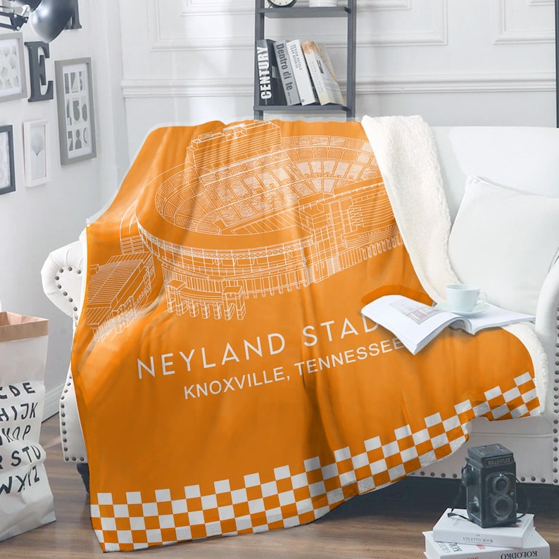 May include: An orange and white stadium blanket featuring a detailed architectural drawing of Neyland Stadium in Knoxville, Tennessee. The blanket has a white checkered border and a soft, white sherpa lining. The blanket is draped over a white couch.