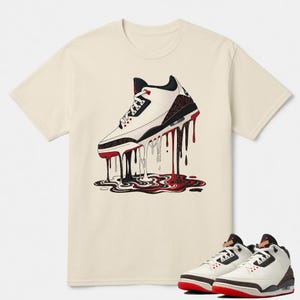 May include: Cream-colored t-shirt featuring a graphic of a white, black, and red sneaker with a dripping paint design. The design includes a pair of matching sneakers at the bottom. The shirt has a crew neck and short sleeves.