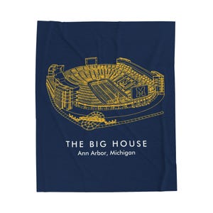 May include: A navy blue blanket featuring a gold line drawing of a stadium. The text "THE BIG HOUSE" and "Ann Arbor, Michigan" are printed in white below the stadium illustration. The blanket is rectangular.