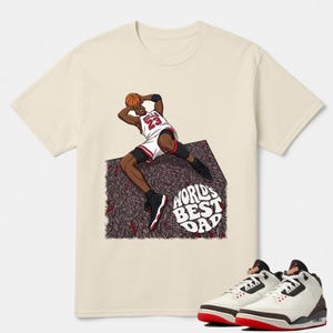May include: Cream-colored t-shirt featuring a graphic of a basketball player in a white and red uniform, holding a basketball. The text "WORLD'S BEST DAD" is below the graphic. A pair of white, red, and brown sneakers are at the bottom.