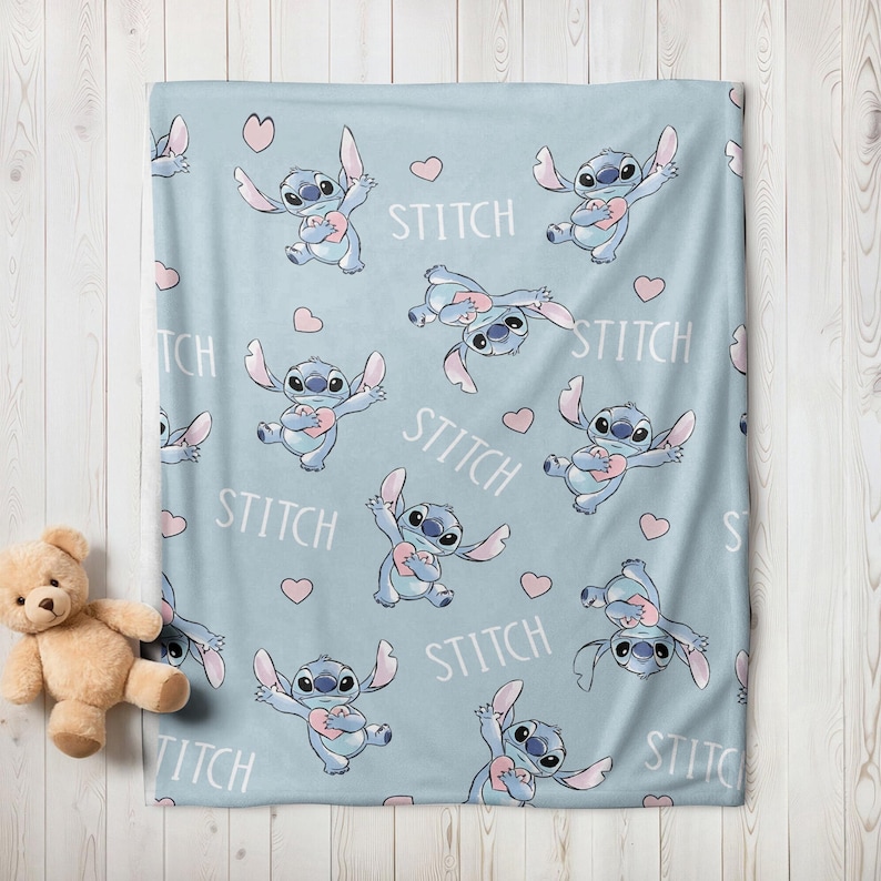 May include: A light blue blanket featuring a pattern of the character Stitch holding a pink heart, with the word "STITCH" in white. Pink hearts are scattered across the blanket. A teddy bear is visible in the corner.