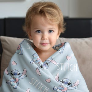 May include: A light blue blanket featuring the character Stitch from the Disney movie Lilo & Stitch. The blanket has a repeating pattern of Stitch, hearts, and the word "STITCH" in white. The blanket is wrapped around a young child.