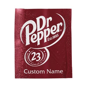 May include: A burgundy blanket featuring the Dr Pepper logo in white, with the text "Est. 1885" and a circular emblem stating "23 Authentic Blend of 23 Flavors." The words "Custom Name" are printed at the bottom.