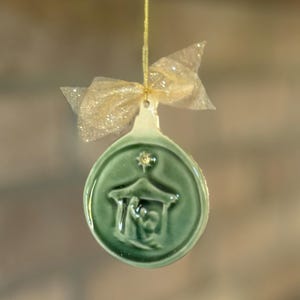 May include: A green ceramic Christmas ornament with a nativity scene design. The ornament is round with a star at the top and a gold bow. The ornament is hanging from a gold string.