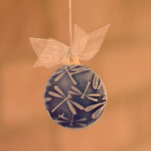 May include: A round, blue and white ceramic ornament with a dragonfly pattern. The ornament is suspended by a white ribbon and topped with a white bow. The ornament is approximately 2 inches in diameter.