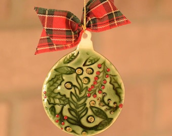 May include: A ceramic Christmas ornament with a green base, decorated with leaves, red berries, and gold accents. A red and green plaid bow is attached to the top with a gold string. The ornament is round and has a glossy finish.