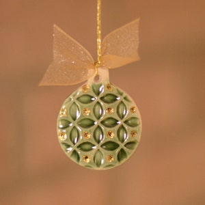 May include: A green ceramic Christmas ornament with a geometric pattern and gold accents. The ornament is round with a small loop at the top, and is suspended by a gold string and a sheer gold bow.