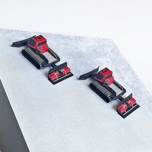 Könnte beinhalten: Zwei rote und schwarze Miniatur-Pistenbullys mit schwarzen Ketten und einem Pflug auf einer weißen Oberfläche.