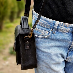 May include: A black leather satchel bag with a long strap worn over the shoulder. The bag has a flap closure and a brass buckle. The bag is worn with blue jeans.
