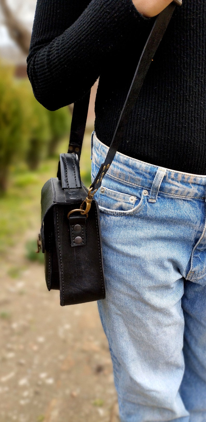 May include: A black leather satchel bag with a long strap worn over the shoulder. The bag has a flap closure and a brass buckle. The bag is worn with blue jeans.