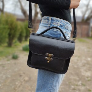 May include: A black leather satchel bag with a brass clasp and a long adjustable strap. The bag is worn over the shoulder of a person wearing blue jeans.