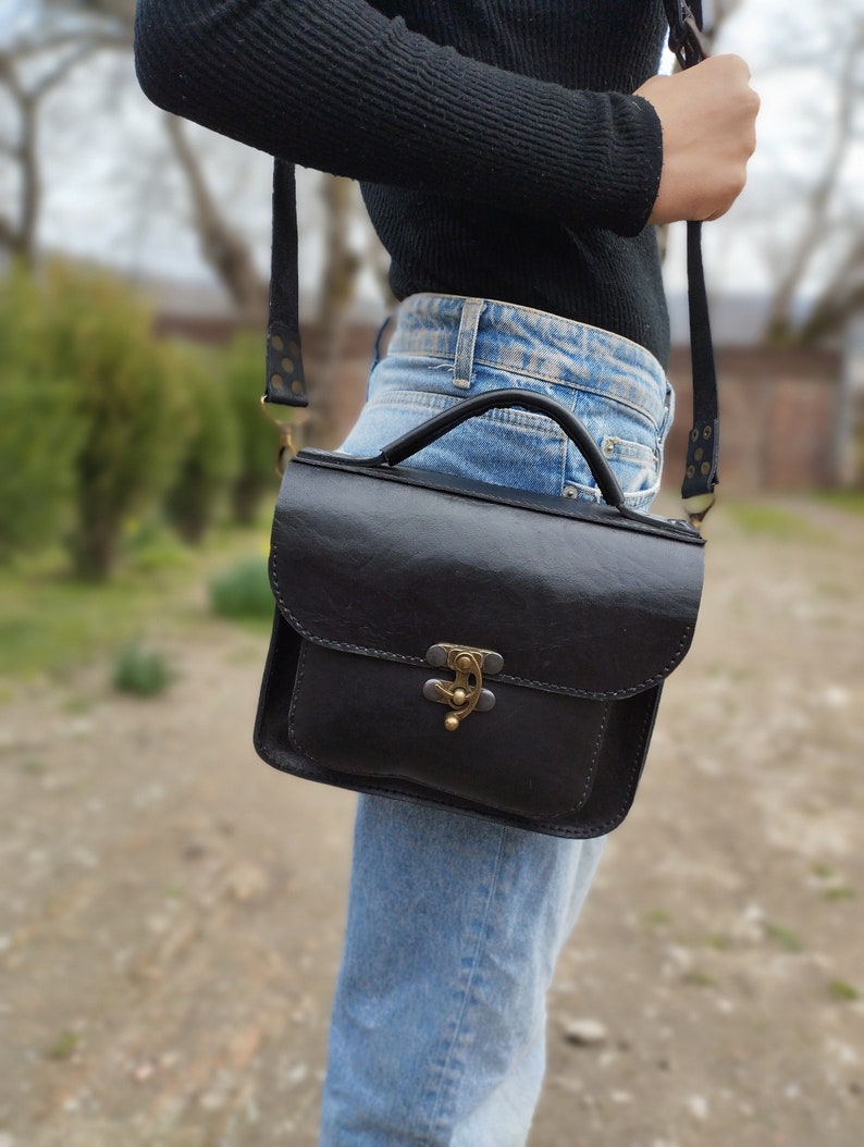 May include: A black leather satchel bag with a brass clasp and a long adjustable strap. The bag is worn over the shoulder of a person wearing blue jeans.