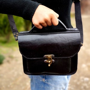 May include: A black leather satchel bag with a brass clasp and a strap. The bag is being held by a person wearing a black sweater and blue jeans.