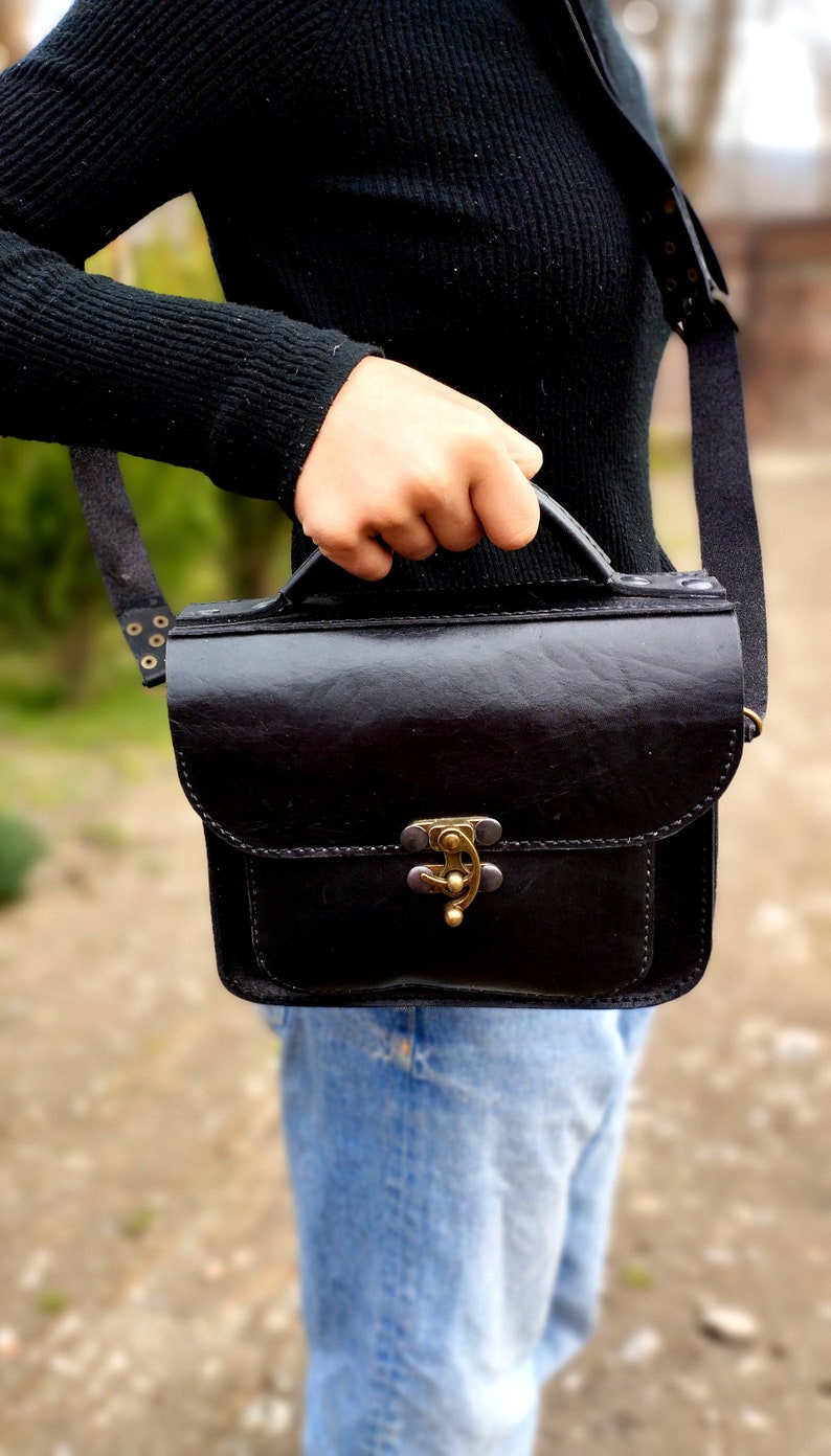 May include: A black leather satchel bag with a brass clasp and a strap. The bag is being held by a person wearing a black sweater and blue jeans.