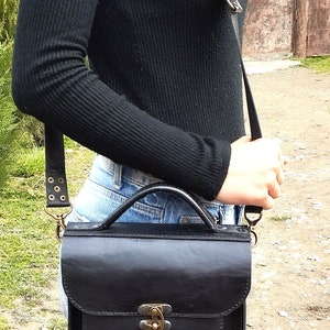 May include: A black leather satchel bag with a brass clasp and a long adjustable strap. The bag is worn over the shoulder of a person wearing a black sweater and blue jeans.