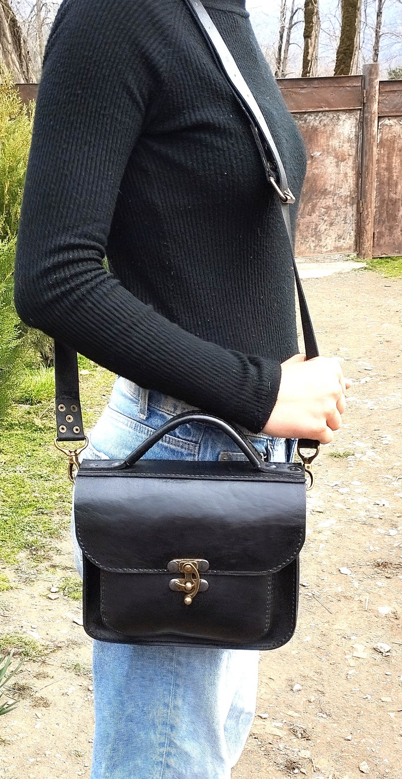 May include: A black leather satchel bag with a brass clasp and a long adjustable strap. The bag is worn over the shoulder of a person wearing a black sweater and blue jeans.