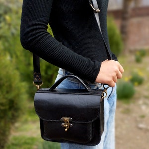 May include: A black leather satchel bag with a brass clasp and a long adjustable strap. The bag is worn over the shoulder of a person wearing blue jeans.