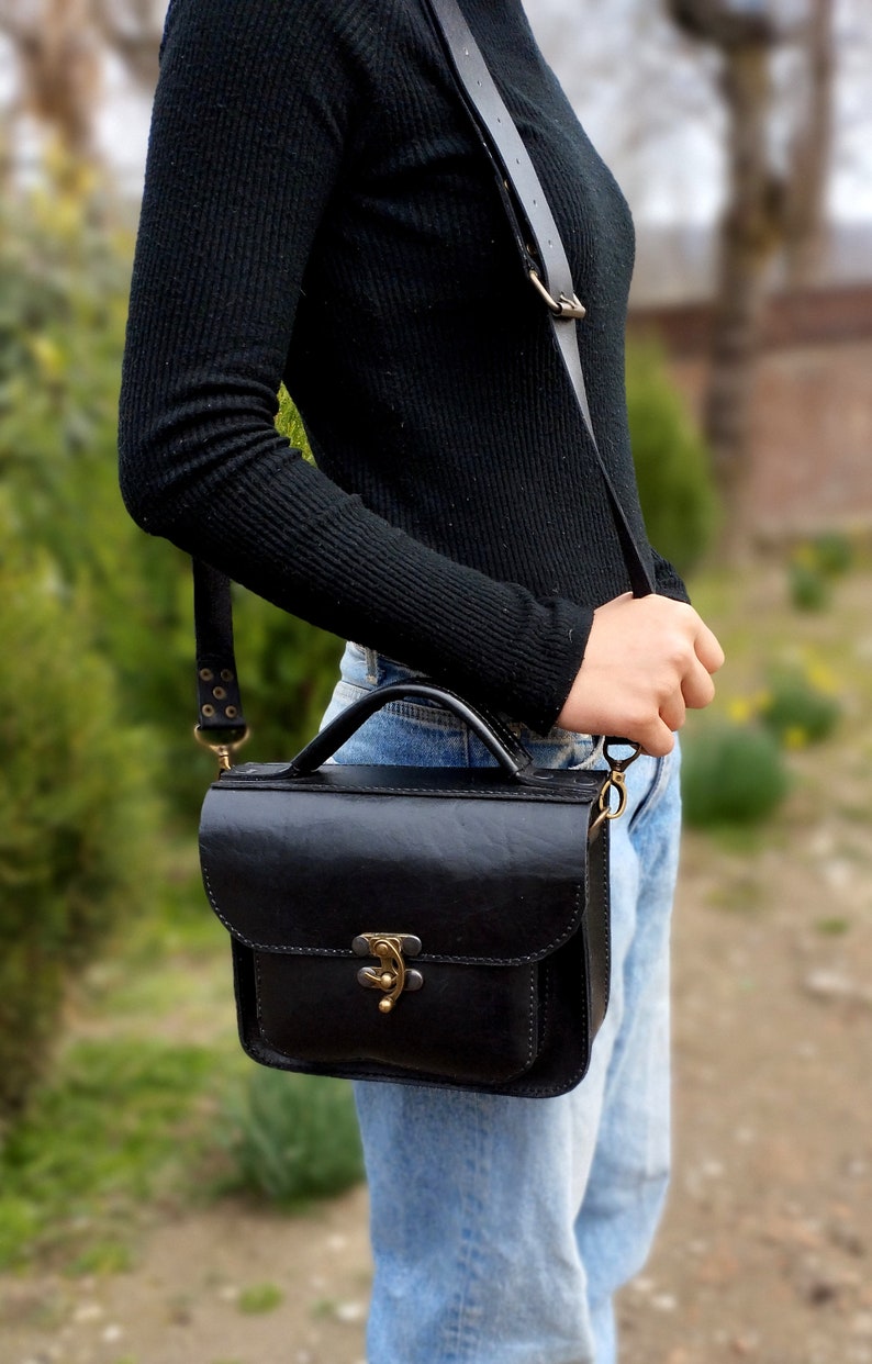 May include: A black leather satchel bag with a brass clasp and a long adjustable strap. The bag is worn over the shoulder of a person wearing blue jeans.