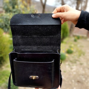 May include: A black leather satchel bag with a flap closure and a small front pocket. The bag has a brass buckle and a long strap.