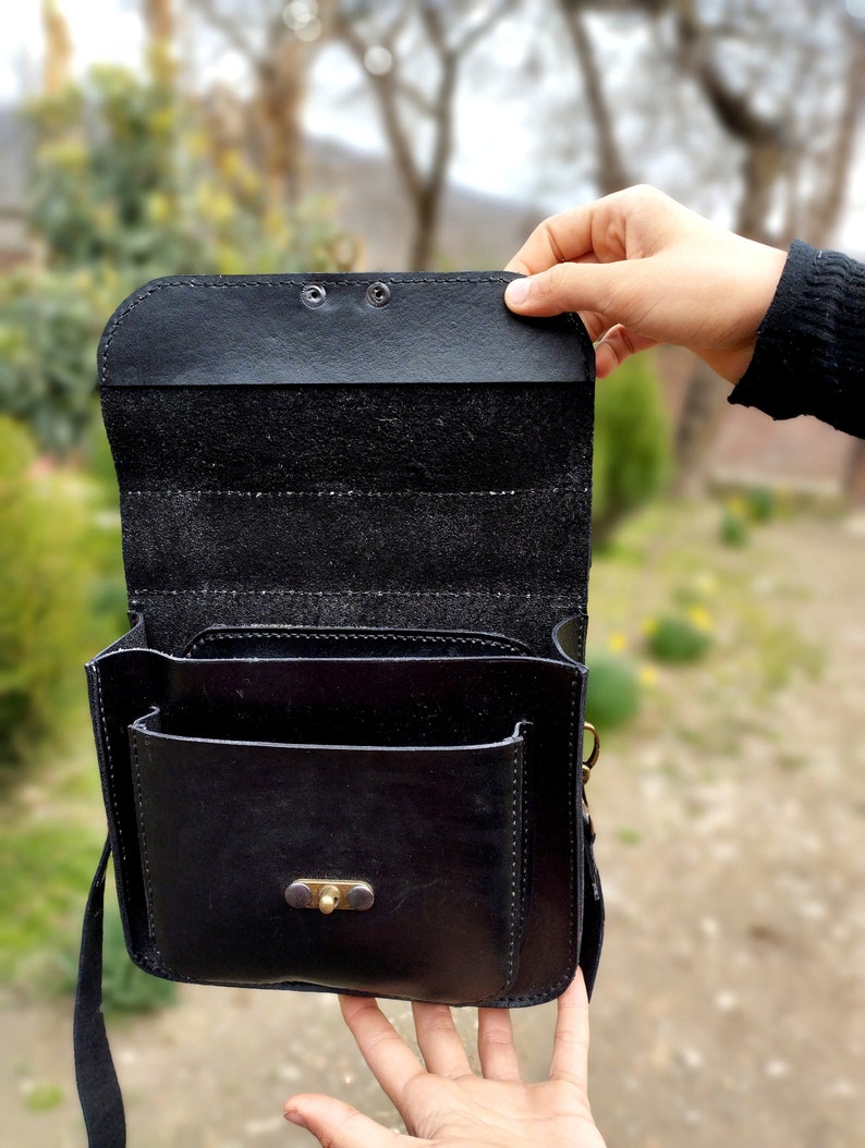 May include: A black leather satchel bag with a flap closure and a small front pocket. The bag has a brass buckle and a long strap.