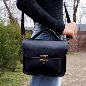 May include: A black leather satchel bag with a top handle and a long adjustable strap. The bag has a brass clasp and a flap closure.