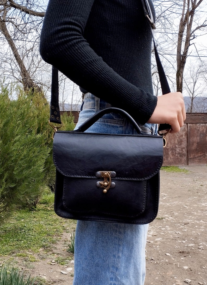 May include: A black leather satchel bag with a top handle and a long adjustable strap. The bag has a brass clasp and a flap closure.