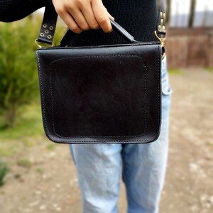 May include: A black leather satchel bag with a top handle and a shoulder strap. The bag has gold-colored hardware and stitching.