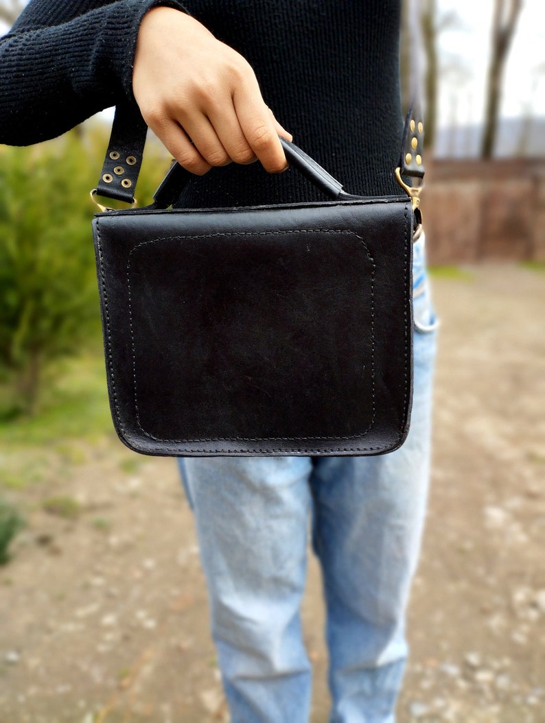 May include: A black leather satchel bag with a top handle and a shoulder strap. The bag has gold-colored hardware and stitching.