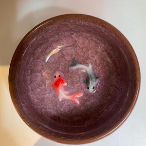 Puede incluir: Un cuenco decorativo de cer&aacute;mica con un esmalte craquelado en tonos morados y marrones. El cuenco contiene tres peque&ntilde;as y detalladas figuras de peces koi en rojo, gris y blanco. El borde del cuenco es marr&oacute;n oscuro.