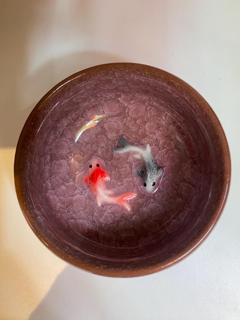 Puede incluir: Un cuenco decorativo de cer&aacute;mica con un esmalte craquelado en tonos morados y marrones. El cuenco contiene tres peque&ntilde;as y detalladas figuras de peces koi en rojo, gris y blanco. El borde del cuenco es marr&oacute;n oscuro.