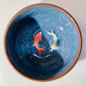Puede incluir: Un cuenco redondo de cer&aacute;mica azul con efecto craquelado. Tiene un borde marr&oacute;n y dos peque&ntilde;as figuras de peces, una roja y otra blanca, en el interior. El cuenco es probablemente decorativo.