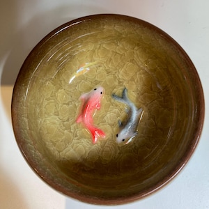 Puede incluir: Un cuenco de cer&aacute;mica con un interior texturizado y agrietado y un borde marr&oacute;n. Dos peque&ntilde;as esculturas de peces detalladas, una roja y otra gris, est&aacute;n colocadas dentro del cuenco, creando una escena acu&aacute;tica en miniatura. El interior del cuenco tiene un tono amarillo-verdoso.