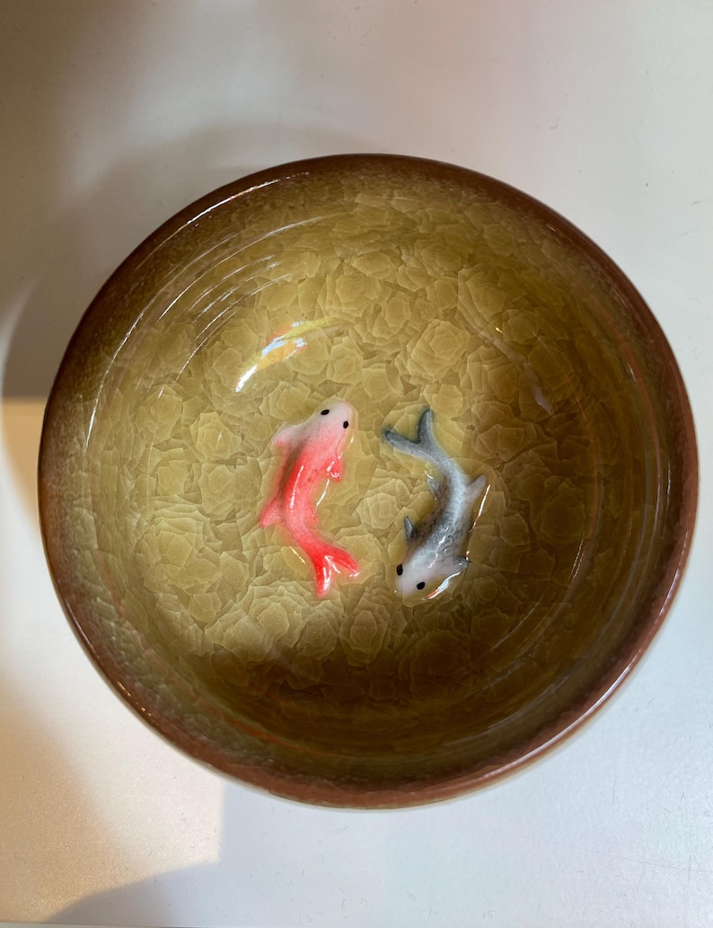 Puede incluir: Un cuenco de cer&aacute;mica con un interior texturizado y agrietado y un borde marr&oacute;n. Dos peque&ntilde;as esculturas de peces detalladas, una roja y otra gris, est&aacute;n colocadas dentro del cuenco, creando una escena acu&aacute;tica en miniatura. El interior del cuenco tiene un tono amarillo-verdoso.