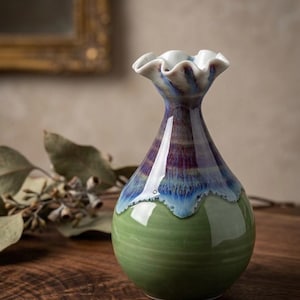 Op de afbeelding: Een handgemaakte keramische vaas met een gerimpelde rand. De vaas heeft een groene basis, die overgaat in blauwe en paarse tinten naar de hals toe, en een witte gerimpelde bovenkant. De vaas wordt gepresenteerd op een houten oppervlak.