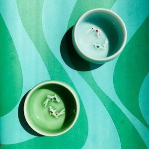May include: Two ceramic bowls, one light blue and one green, each filled with a liquid and featuring small, white koi fish with red and black markings. The bowls sit on a teal and green wavy patterned surface.