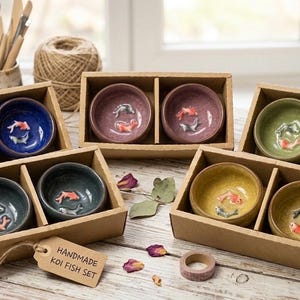May include: Four sets of two small ceramic bowls in brown cardboard boxes. Each bowl features a raised koi fish figure at the bottom. The bowls come in blue, purple, green, and yellow glaze colours. A tag reads HANDMADE KOI FISH SET.