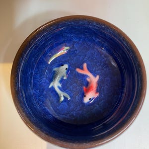 Puede incluir: Un cuenco redondo de cer&aacute;mica azul con efecto craquelado. Dos peces peque&ntilde;os, uno plateado y otro rojo, nadan en el cuenco. El borde del cuenco es de color marr&oacute;n oscuro.