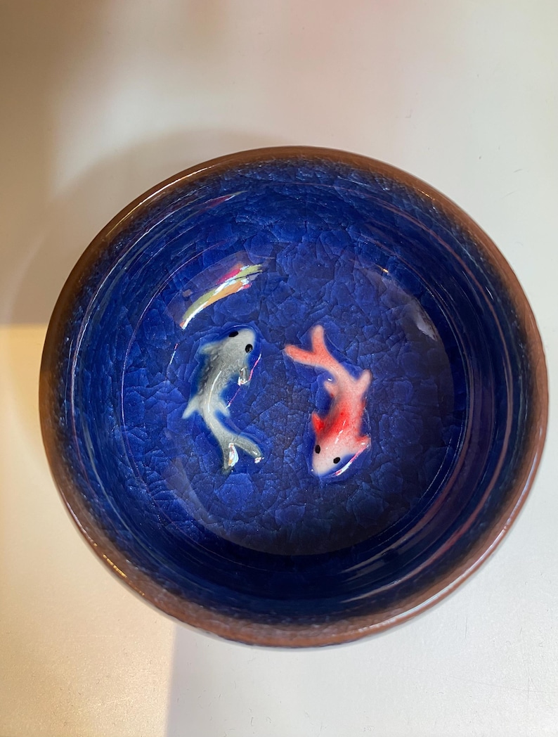 Puede incluir: Un cuenco redondo de cer&aacute;mica azul con efecto craquelado. Dos peces peque&ntilde;os, uno plateado y otro rojo, nadan en el cuenco. El borde del cuenco es de color marr&oacute;n oscuro.