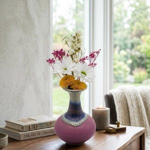 Puede incluir: Un jarrón de cerámica con base rosa y cuello azul y blanco contiene un ramo de margaritas blancas, crisantemos amarillos y flores rosas. El jarrón está sobre una mesa de madera, junto a libros y una vela encendida.