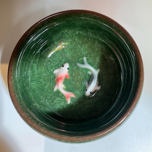 Puede incluir: Un cuenco de cer&aacute;mica verde con efecto craquelado. Dos peque&ntilde;os peces koi de cer&aacute;mica nadan en su interior. Uno es blanco con detalles rojos, el otro gris y blanco. El cuenco tiene un borde marr&oacute;n.