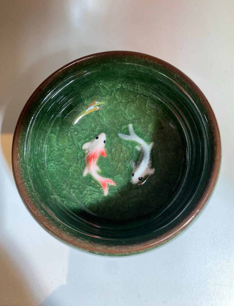 Puede incluir: Un cuenco de cer&aacute;mica verde con efecto craquelado. Dos peque&ntilde;os peces koi de cer&aacute;mica nadan en su interior. Uno es blanco con detalles rojos, el otro gris y blanco. El cuenco tiene un borde marr&oacute;n.