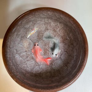 Puede incluir: Un cuenco de cer&aacute;mica con borde marr&oacute;n e interior texturizado. Dos peces peque&ntilde;os, uno rojo y otro gris, est&aacute;n representados dentro del cuenco. El interior del cuenco tiene un patr&oacute;n claro y texturizado, y los peces tienen detalles.