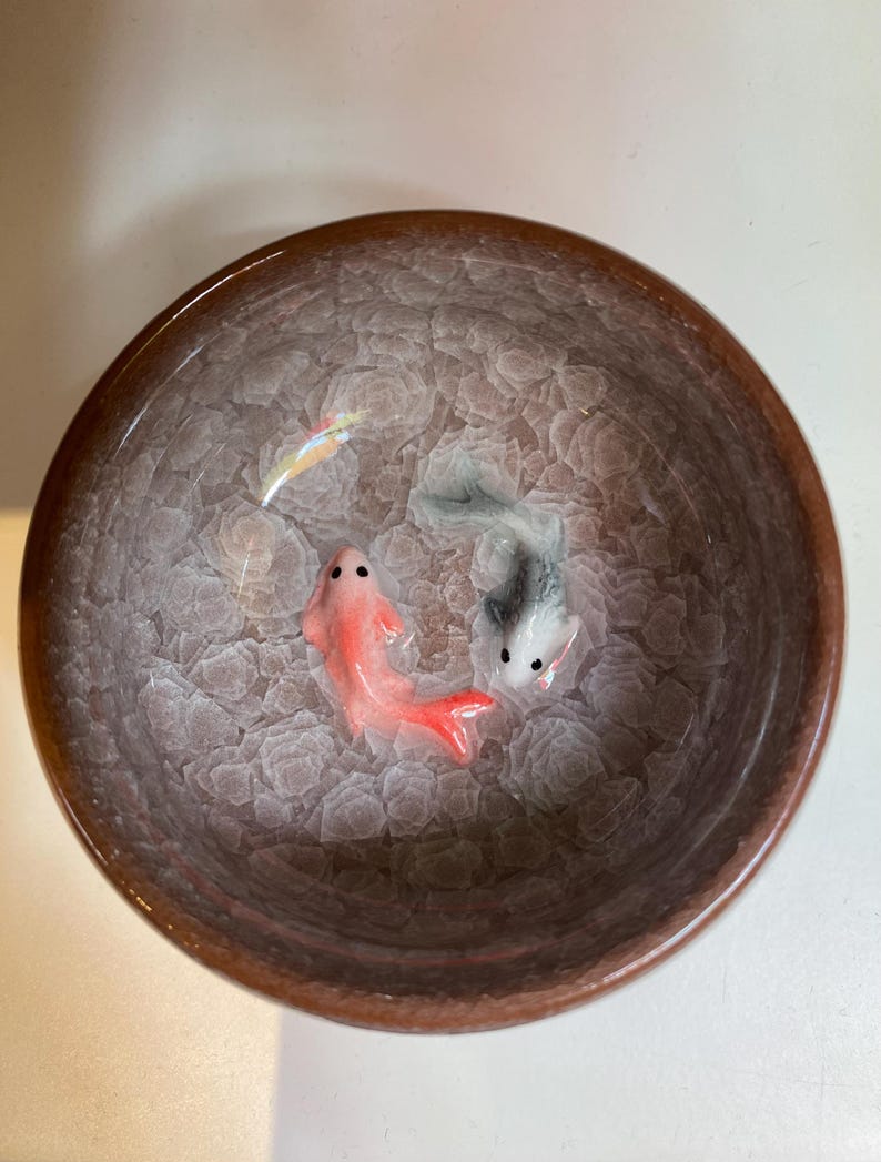 Puede incluir: Un cuenco de cer&aacute;mica con borde marr&oacute;n e interior texturizado. Dos peces peque&ntilde;os, uno rojo y otro gris, est&aacute;n representados dentro del cuenco. El interior del cuenco tiene un patr&oacute;n claro y texturizado, y los peces tienen detalles.
