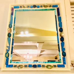 May include: A white framed mirror with a blue and green mosaic tile border. The tiles are various shapes and sizes and include seashells.