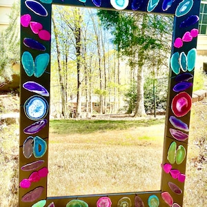 May include: A rectangular mirror with a black frame, adorned with colorful, irregularly shaped agate slices in shades of blue, purple, pink, and green. The mirror is outdoors, reflecting a natural landscape.