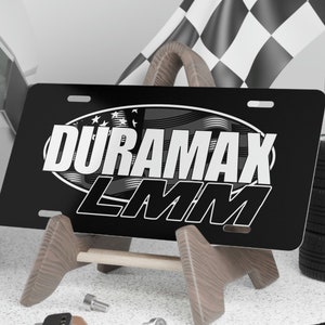 May include: Black and silver Duramax LMM license plate with a wood stand. The license plate has a red, white, and blue American flag design.